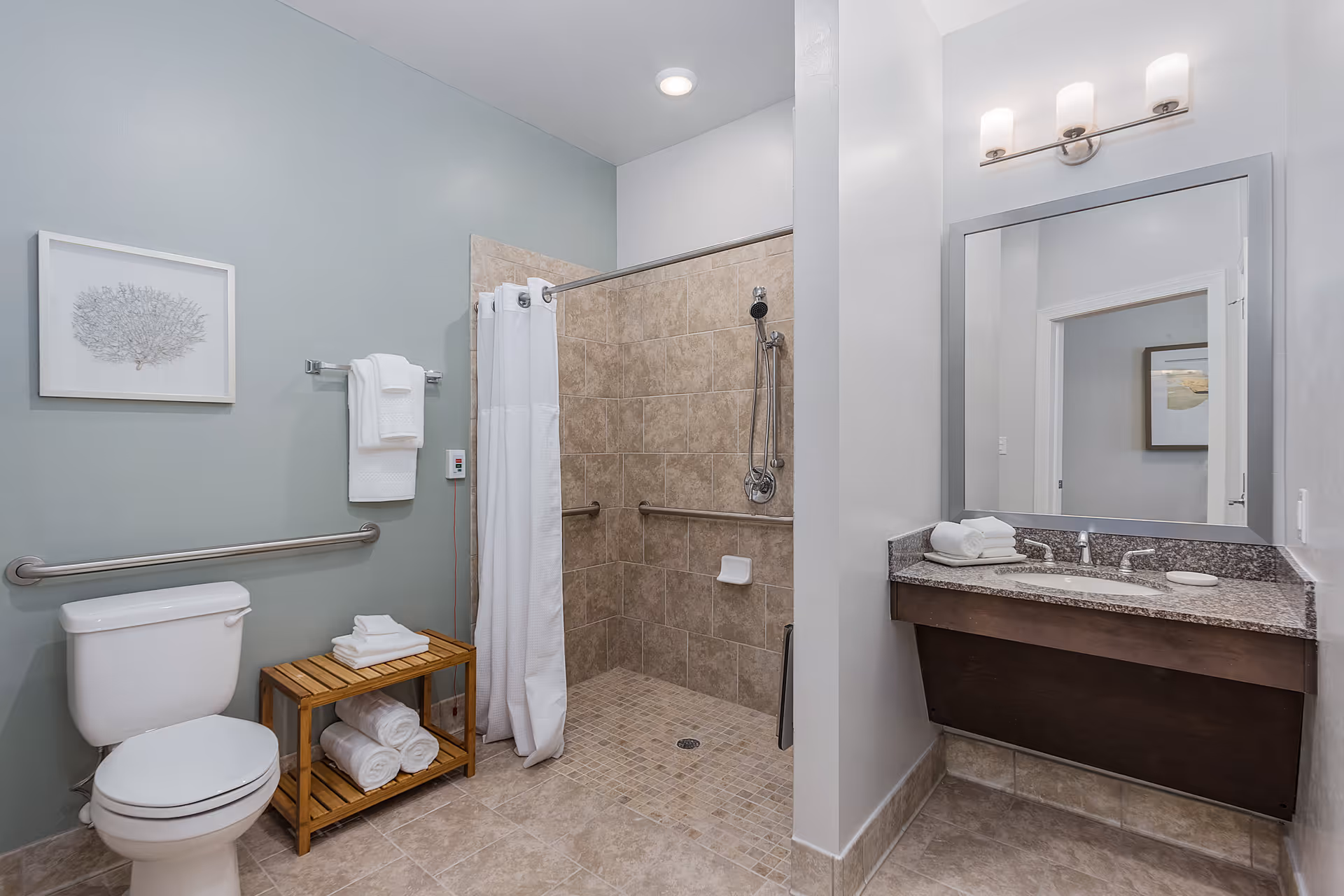 A clean and modern bathroom featuring a white toilet, a wooden shelf with rolled towels, a walk-in tiled shower with a white curtain and grab bars, and a vanity with a granite countertop, sink, mirror, and light fixture above.