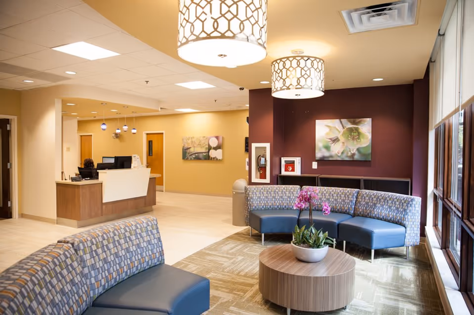 A bright and welcoming hospital lobby area with patterned blue and gray curved seating around a round wooden table with a potted orchid plant. The reception desk is visible in the background with a staff member seated. The walls are painted in warm beige and maroon tones, decorated with floral artwork. Large windows on the right let in natural light, and modern pendant lights hang from the ceiling.