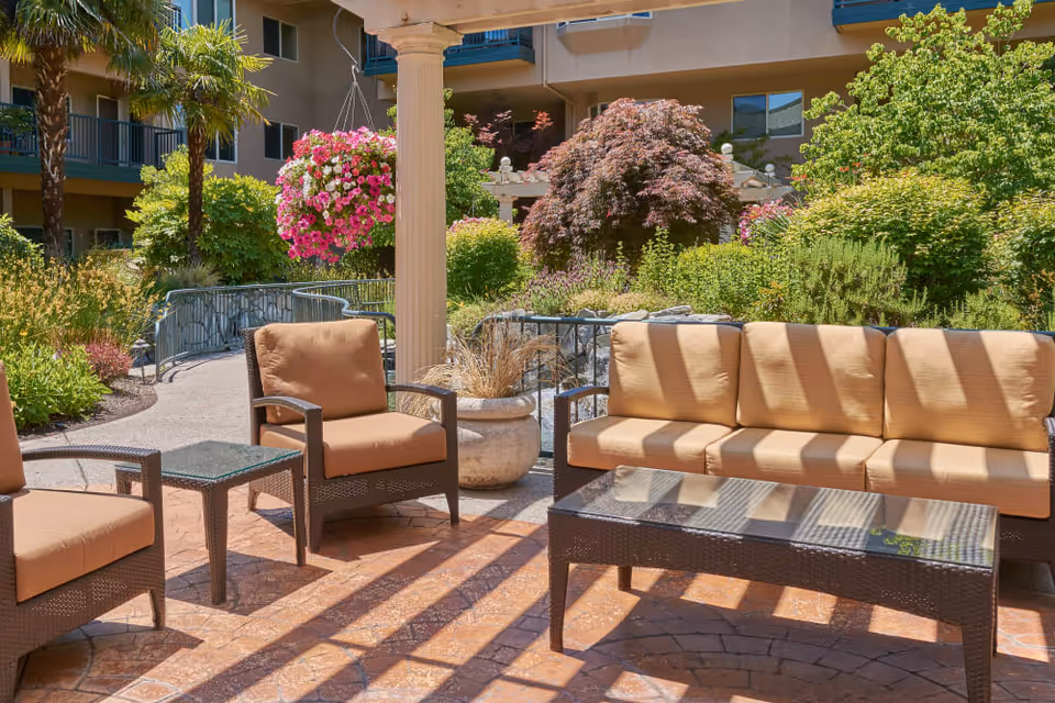 Outdoor patio area with cushioned wicker chairs and a sofa around a glass-top coffee table, surrounded by lush greenery, flowering plants, and a building in the background.