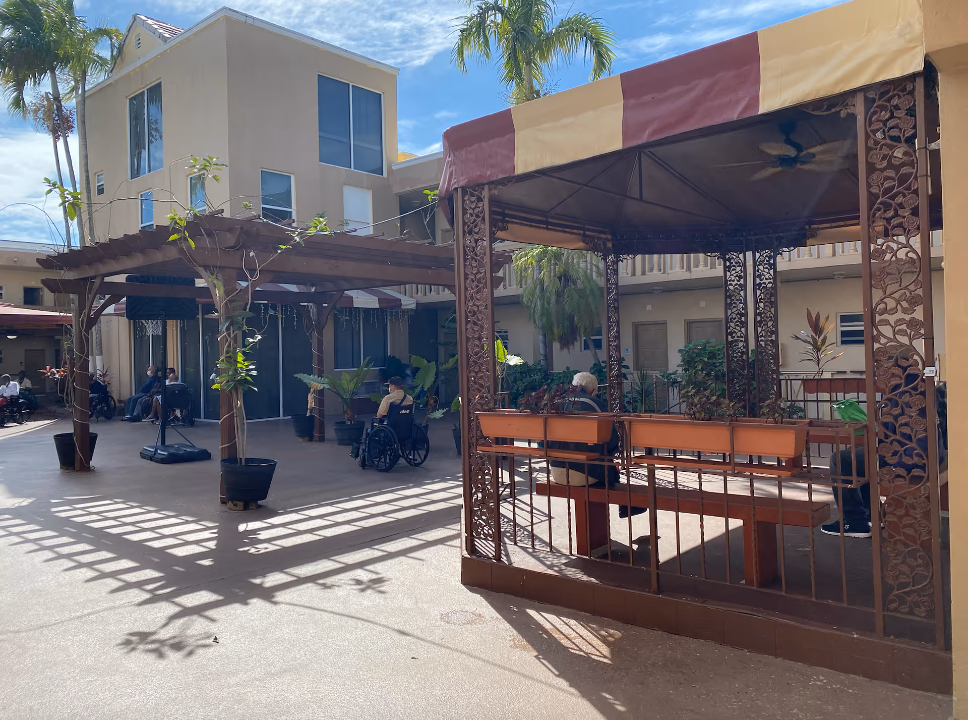 Outdoor courtyard area at Courtyard Plaza with a gazebo featuring decorative metalwork and benches. Several elderly individuals are seated, some in wheelchairs, enjoying the sunny day. The courtyard is surrounded by a two-story building with windows and doors, and there are potted plants and palm trees around.