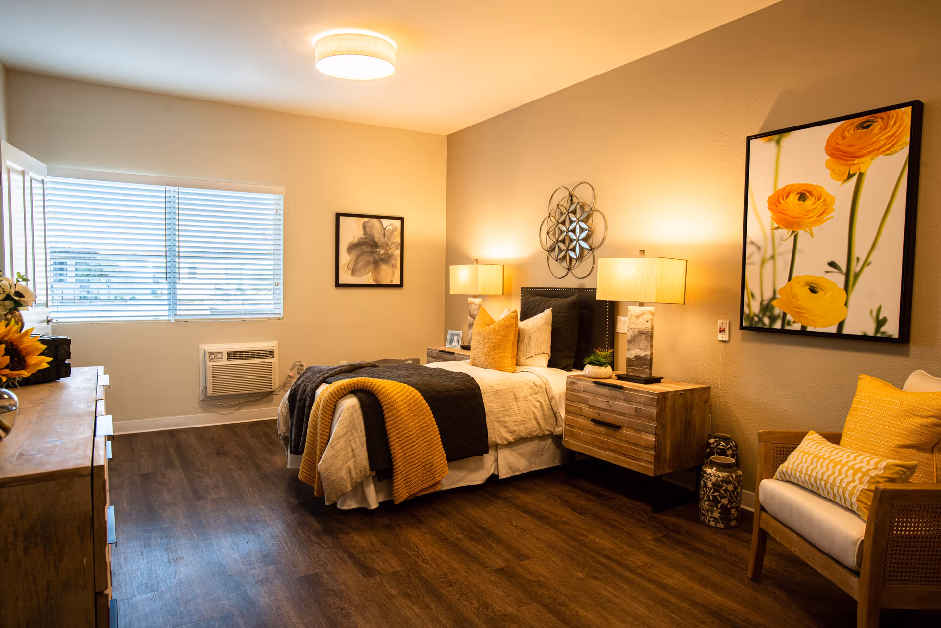 A cozy bedroom with a neatly made bed featuring yellow and gray bedding. There are two wooden nightstands with lamps on either side of the bed. The room has wooden flooring, a window with blinds, a dresser with flowers, and two framed artworks on the walls. A comfortable chair with yellow cushions is placed near the bed.