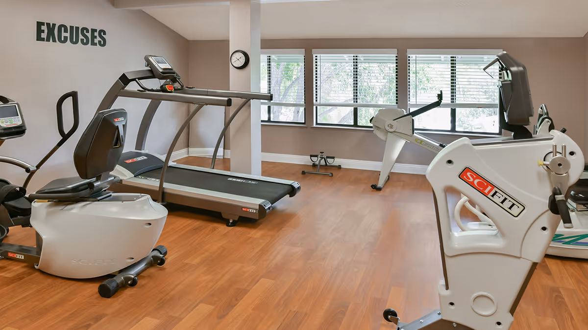 Exercise room with treadmills, stationary bikes, and a rowing machine on a wood floor beneath windows.