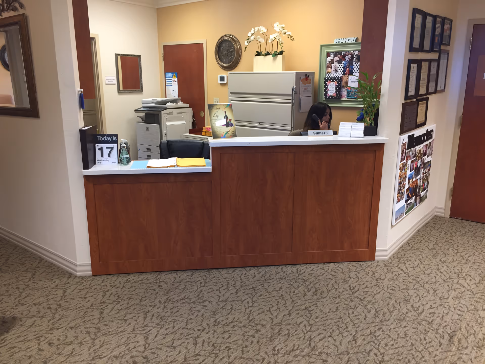Reception desk area in an office with a woman sitting behind the desk talking on the phone. The desk has a nameplate with the name Sumera, a calendar showing the date July 17, 2019, and some office supplies. Behind the desk are filing cabinets, a clock on the wall, and a bulletin board with photos and notes. The walls are painted beige and light yellow, and the floor is carpeted with a patterned design.