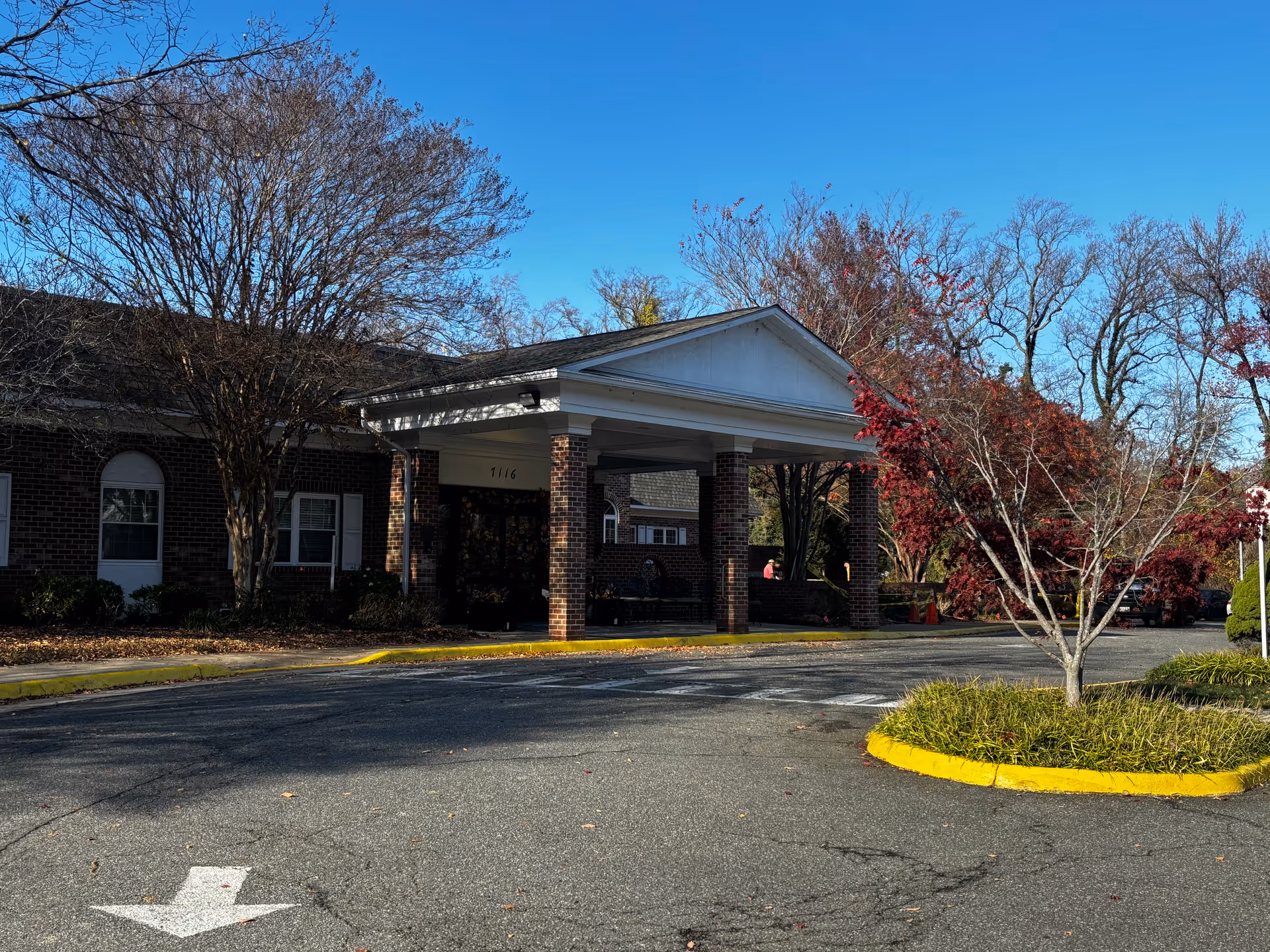 Driveway and covered entrance/portico of a brick senior living building with trees under a clear blue sky.