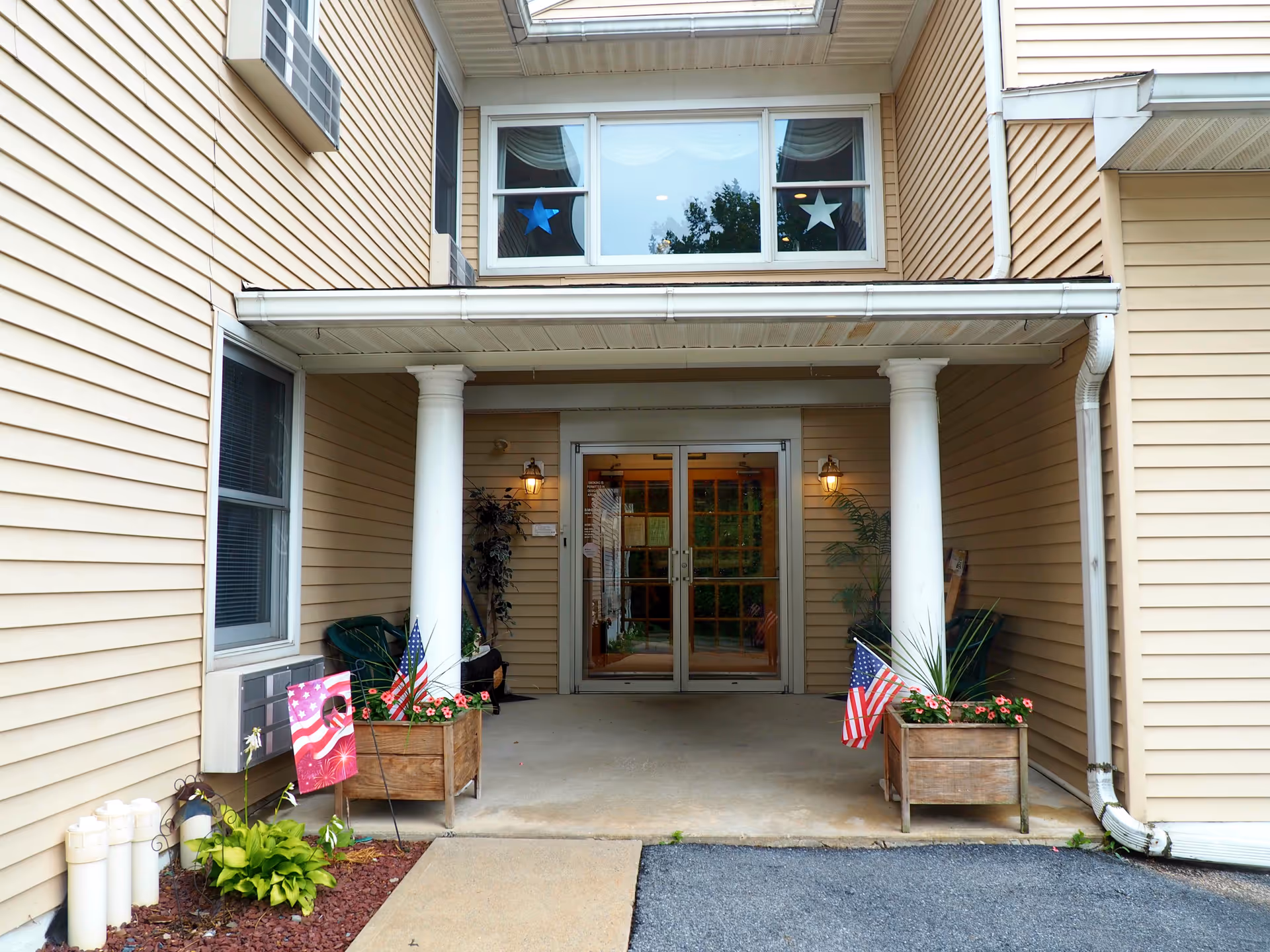 Entrance to a building with beige siding, two white columns supporting a small roof over the doorway, and double glass doors. There are two wooden planters with pink flowers and small American flags on either side of the entrance. Above the door is a window with blue and white star decorations.