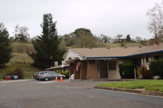 Single-story healthcare center with a covered entrance, American flag, parked cars, and grassy hillside in the background.