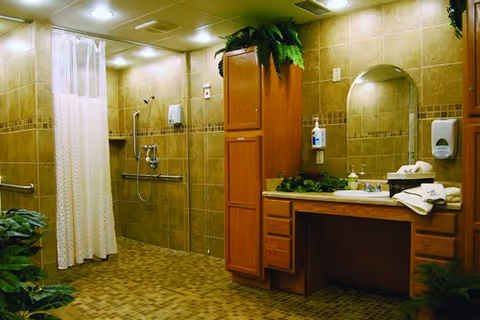 A spacious bathroom with tan tiled walls and floor, featuring a walk-in shower area with a white curtain, grab bars, and a handheld showerhead. There is a wooden vanity with a large mirror, a sink, soap dispenser, and neatly folded towels. Green plants are placed on top of the cabinets and around the room, adding a touch of nature.