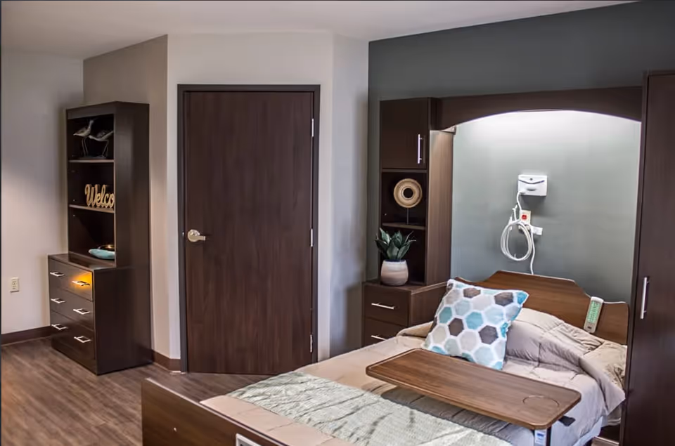A furnished patient bedroom with a hospital-style bed, overbed table, headboard cabinets, and a closed wooden door.