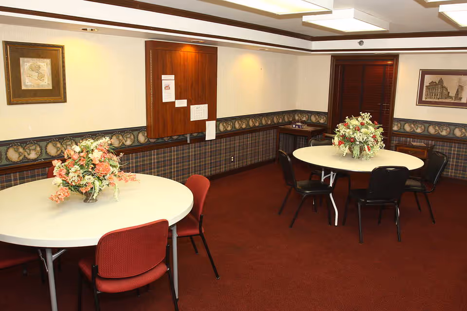 A small dining room with two round tables, each decorated with a floral centerpiece. The room has red carpet, patterned wallpaper with a border, framed pictures on the walls, and a wooden cabinet with papers attached. There are red and black chairs around the tables.