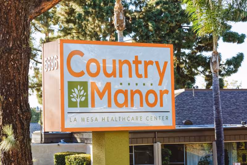 Country Manor sign for La Mesa Healthcare Center beside trees and a building.