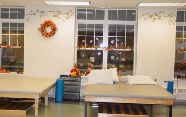 A room with two padded therapy or massage tables, each with white pillows. The room has large windows decorated with autumn-themed stickers and garlands. A fall wreath hangs on the wall between the windows. There is a small black storage unit between the tables and a blue cylindrical bolster next to each table. The walls are light-colored with a floral vine border near the ceiling.