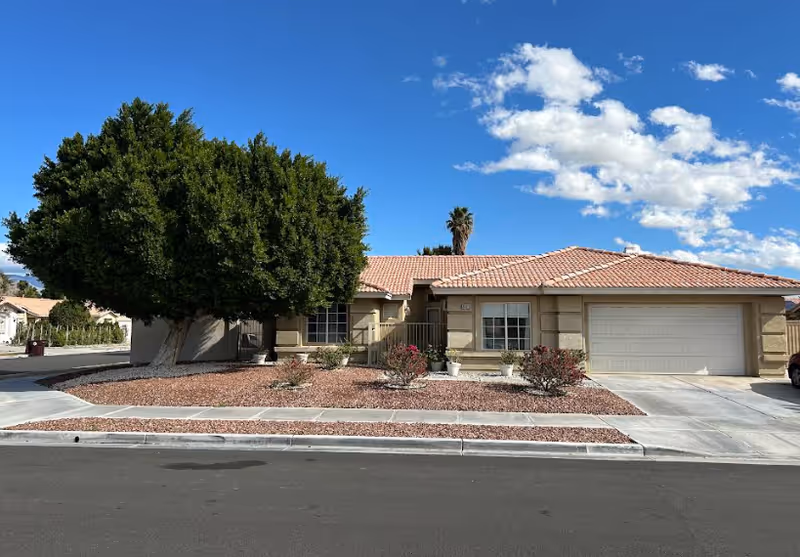 Single-story residential building with a tiled roof, a large tree on the left side, desert landscaping with rocks and small bushes in front, and a two-car garage on the right side under a blue sky with scattered clouds.