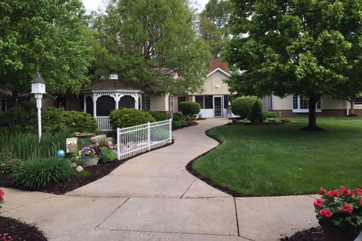 A paved walkway curves through a well-maintained garden area with green grass, trees, and flowering plants. To the left, there is a white gazebo surrounded by bushes and a white birdhouse on a post. In the background, a beige building with a red roof and large windows is visible.