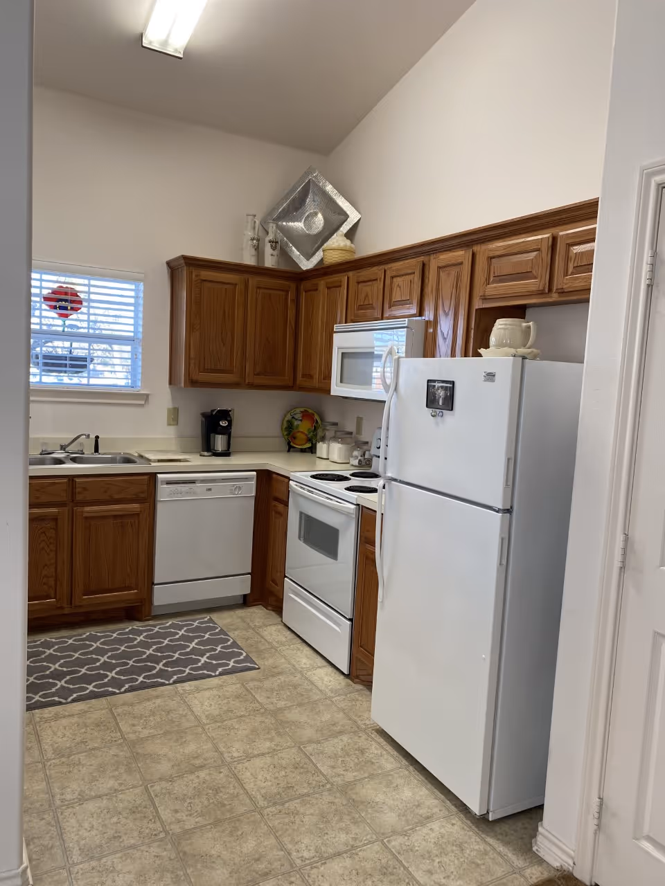A kitchen with wooden cabinets, a white refrigerator, white stove, white microwave, dishwasher, and a double sink. There is a small window with blinds, a coffee maker on the counter, and decorative items on top of the cabinets. The floor is tiled and there is a gray patterned rug in front of the sink.