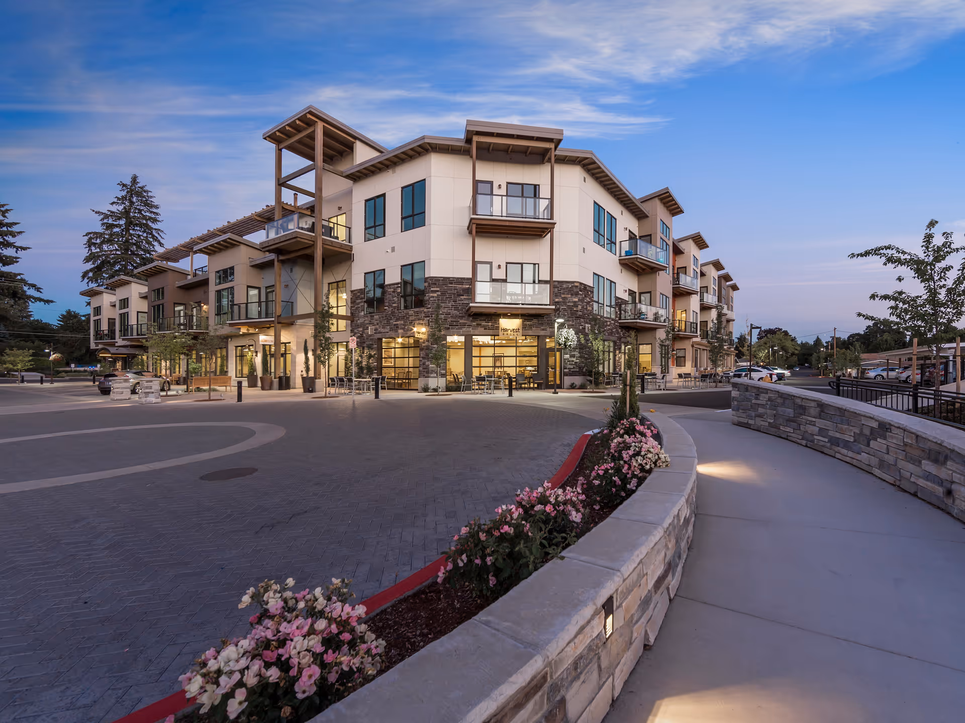 Exterior view of a modern three-story senior living community building at dusk with large windows, balconies, outdoor seating, landscaped flower beds, and a curved stone wall along a sidewalk.