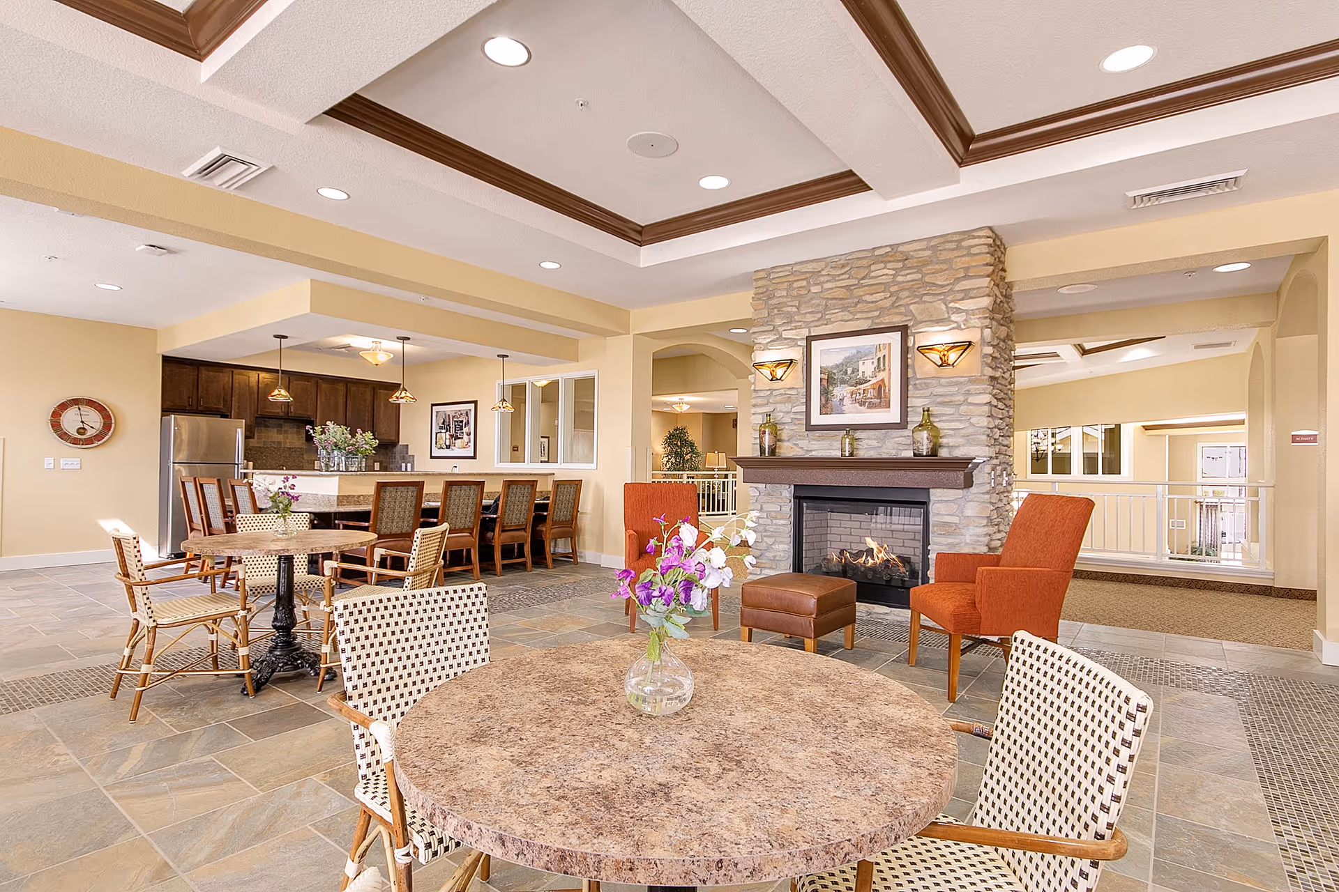 A spacious and well-lit common area featuring a stone fireplace with a framed painting above it, two orange armchairs, and a brown ottoman. In the foreground, there is a round table with a vase of purple and white flowers and woven chairs around it. In the background, a kitchen area with a refrigerator, dark wood cabinets, and a long counter with several chairs is visible. The room has a tiled floor and a tray ceiling with recessed lighting.