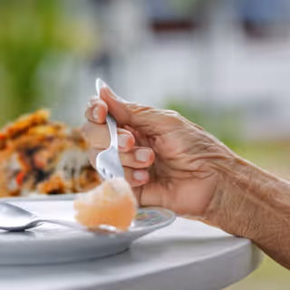 Close-up of an elderly hand holding a fork over a plate at a dining table.