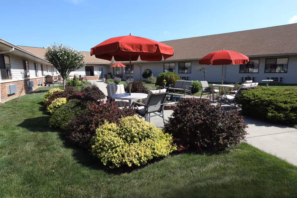 Outdoor courtyard area at The Laurels of Defiance featuring green grass, various bushes, and shrubs. There are several patio tables with red umbrellas and cushioned chairs arranged on a concrete patio. The building surrounds the courtyard with windows and a clear blue sky overhead.