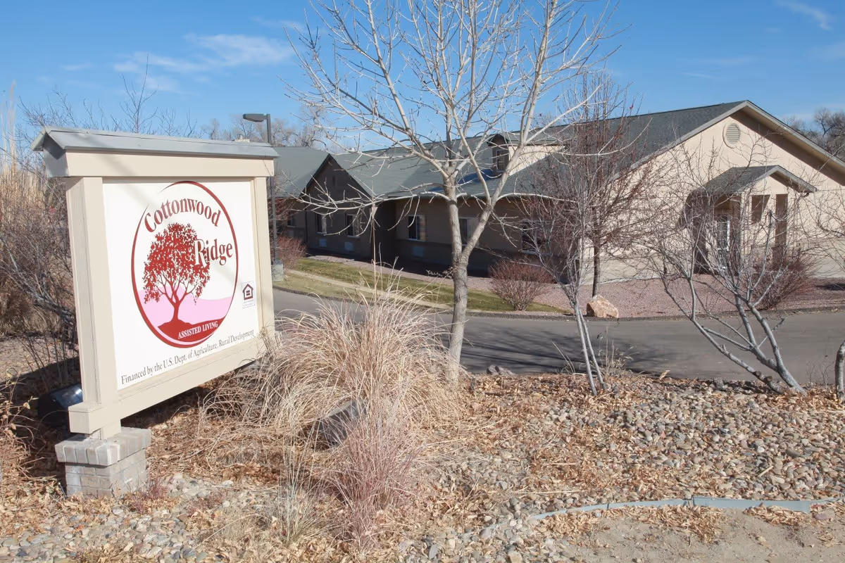 Outdoor view of Cottonwood Ridge assisted living facility with a large sign displaying the facility name and logo in front of a beige building with a green roof, surrounded by leafless trees and dry landscaping under a clear blue sky.