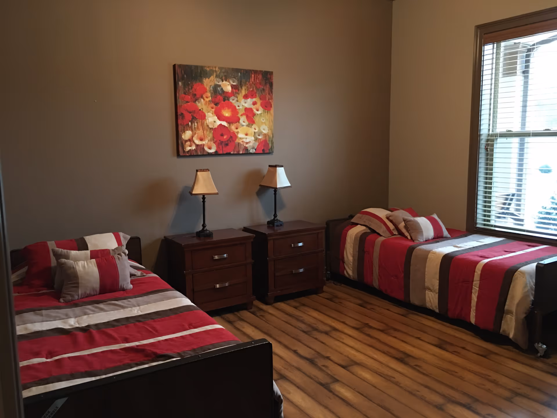 A bedroom with two single beds featuring red, white, and beige striped bedding. Between the beds are two wooden nightstands, each with a lamp. A colorful floral painting hangs on the wall above the nightstands. There is a window with blinds on the right side of the room, and the floor has a wood-like finish.