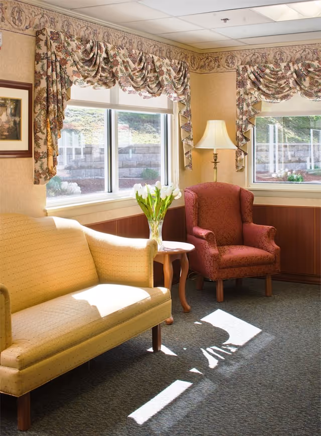A cozy sitting area with a yellow upholstered loveseat and a red patterned armchair. Between them is a small wooden side table holding a vase of white tulips. Two windows with floral valances let in sunlight, casting shadows on the carpeted floor. A floor lamp stands beside the armchair.
