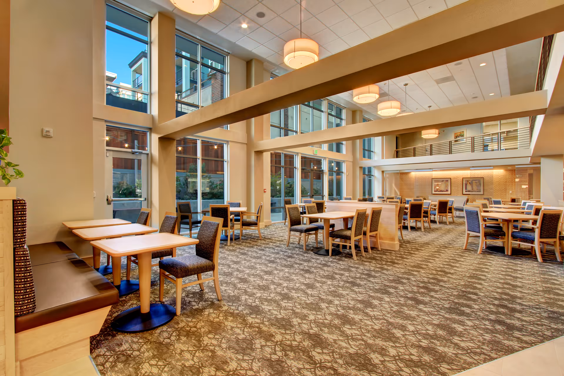 A spacious dining room with multiple tables and chairs arranged neatly. The room features large floor-to-ceiling windows allowing natural light to fill the space. There are modern light fixtures hanging from the ceiling and a patterned carpet covering the floor. The walls are painted in neutral tones, and there is a balcony or mezzanine level visible above.