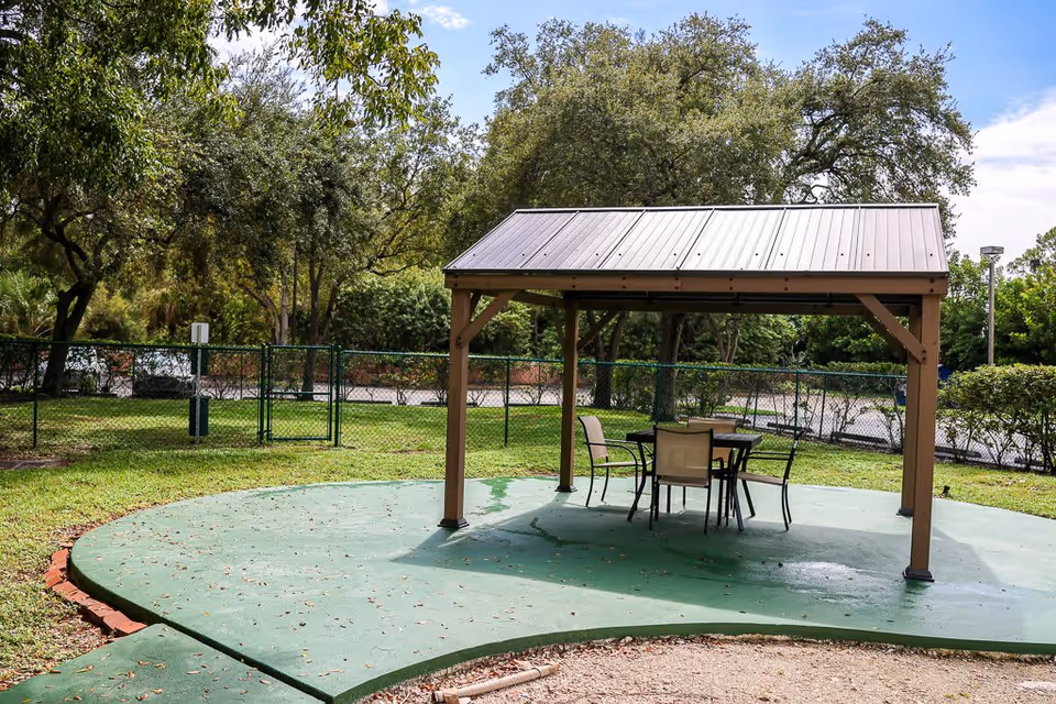 Outdoor seating area with a small metal roof structure covering a table and four chairs, surrounded by green grass, trees, and a chain-link fence in the background under a partly cloudy sky.
