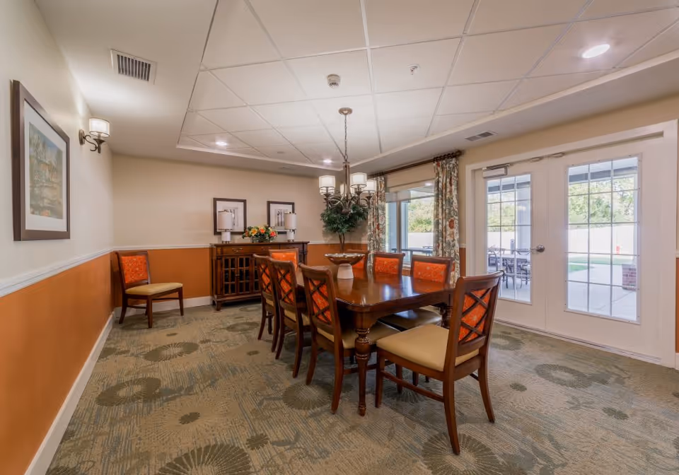 A dining room with a wooden table surrounded by eight chairs, six with orange patterned cushions and two with beige cushions. The room has a patterned carpet, a sideboard with lamps and flowers, framed pictures on the walls, and a chandelier hanging above the table. There are large glass doors leading to an outdoor patio area with tables and chairs. The walls are painted beige with an orange accent below the chair rail.