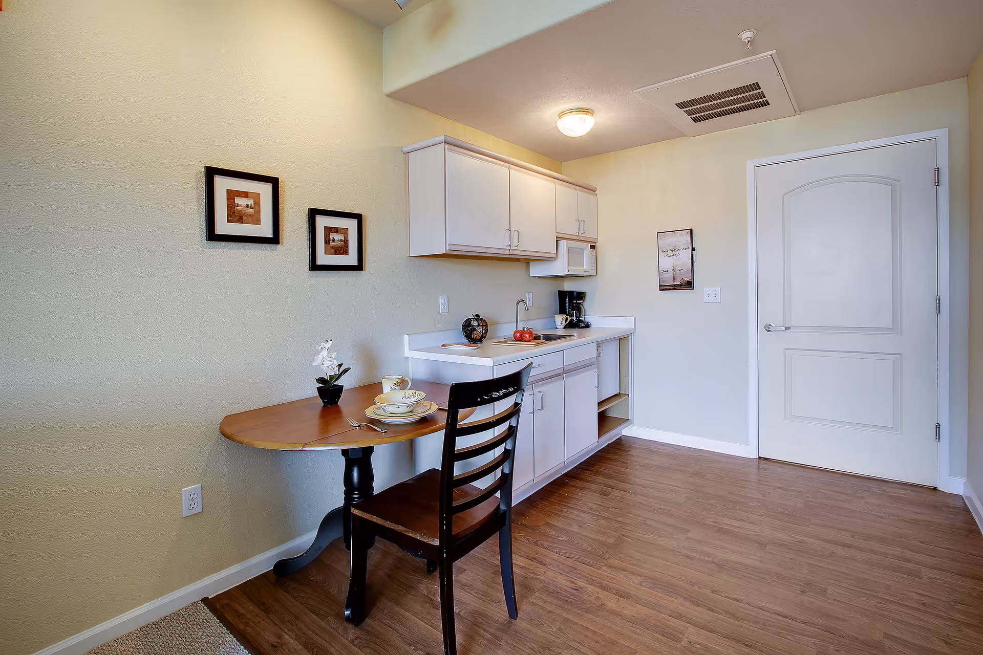 A small kitchen area with white cabinets, a microwave, a coffee maker, and a sink. There is a wooden table with a single chair, set with a cup, saucer, plate, and fork. Two framed pictures hang on the wall above the table, and a closed white door is visible on the right side. The floor is wood, and the walls are painted light yellow.