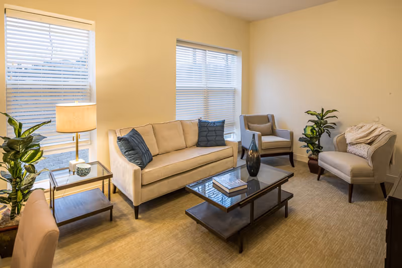Bright, furnished living room with a beige sofa, two armchairs, a glass-top coffee table, side tables, lamps, and potted plants.