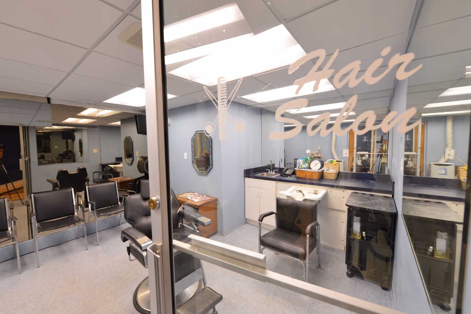 Interior view of a hair salon with multiple black salon chairs, mirrors on the walls, a counter with a sink, and various hair care products. The glass door has the words 'Hair Salon' along with icons of scissors and a comb.