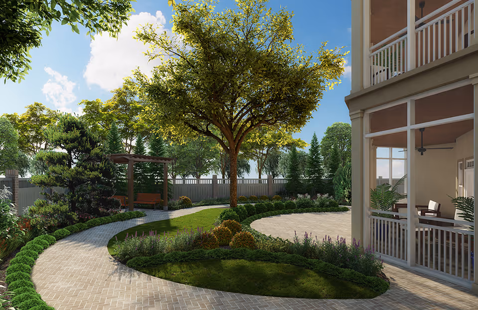 A landscaped outdoor garden area with a curved paved walkway, green grass, various shrubs, and a large tree in the center. There is a wooden pergola with a bench underneath it in the background, and a building with a covered porch featuring chairs and plants on the right side. The sky is blue with some clouds.