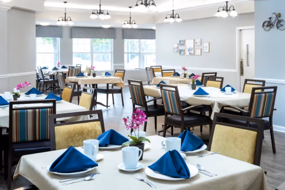 A bright dining room with multiple tables covered in beige tablecloths, each set with white plates, blue folded napkins, white cups, and silverware. The chairs have striped and solid yellow upholstery. Pink flowers in small pots decorate the tables. Large windows with gray valances allow natural light to fill the room. The walls are light gray with white trim, and there are framed pictures and a decorative bicycle on the walls.