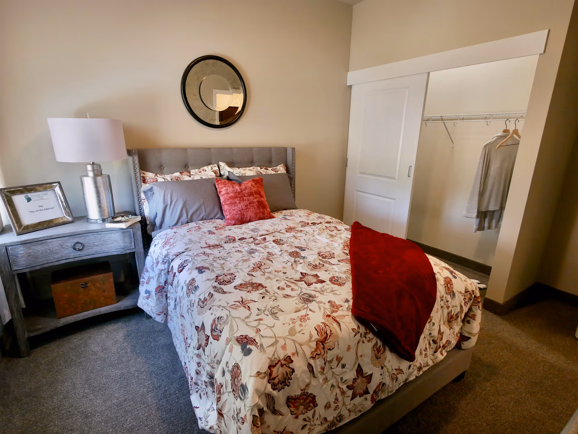 Furnished bedroom with a floral bedspread, gray upholstered headboard, nightstand with a lamp, and an open closet with sliding door.