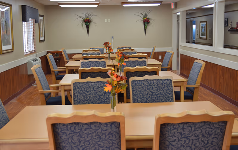 Dining room with rows of wooden tables and upholstered chairs, each table decorated with a small floral centerpiece.