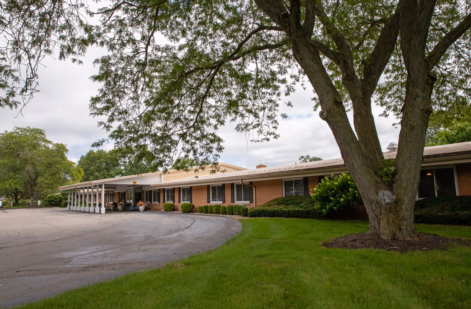 Exterior view of a single-story brick building with a covered entrance and a curved driveway. There is a large tree with green leaves in the foreground and well-maintained grass and shrubs surrounding the building under a cloudy sky.