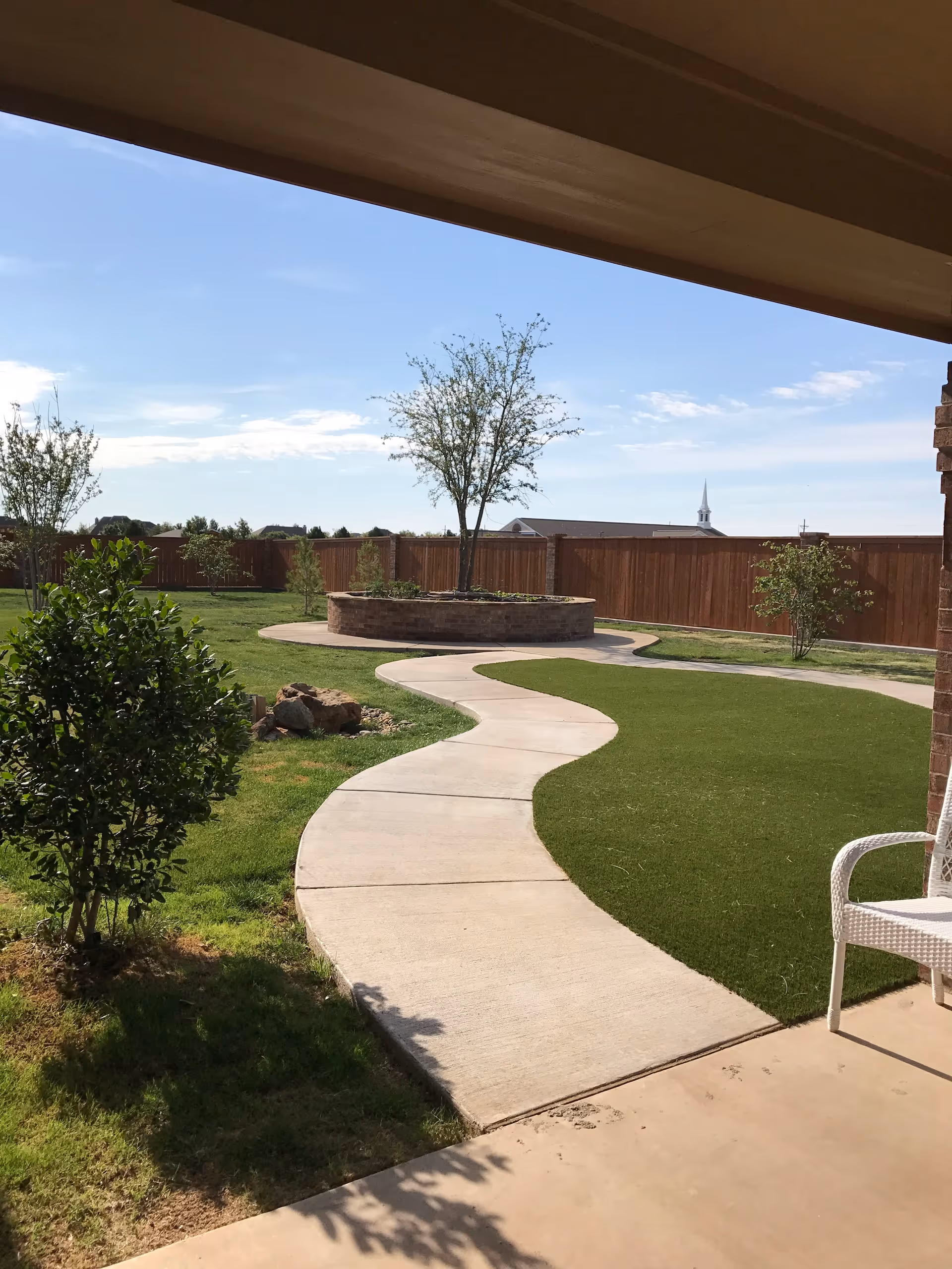 Curving concrete walkway through a fenced sunny courtyard with trees, grass, and a raised circular planter seen from a covered patio.