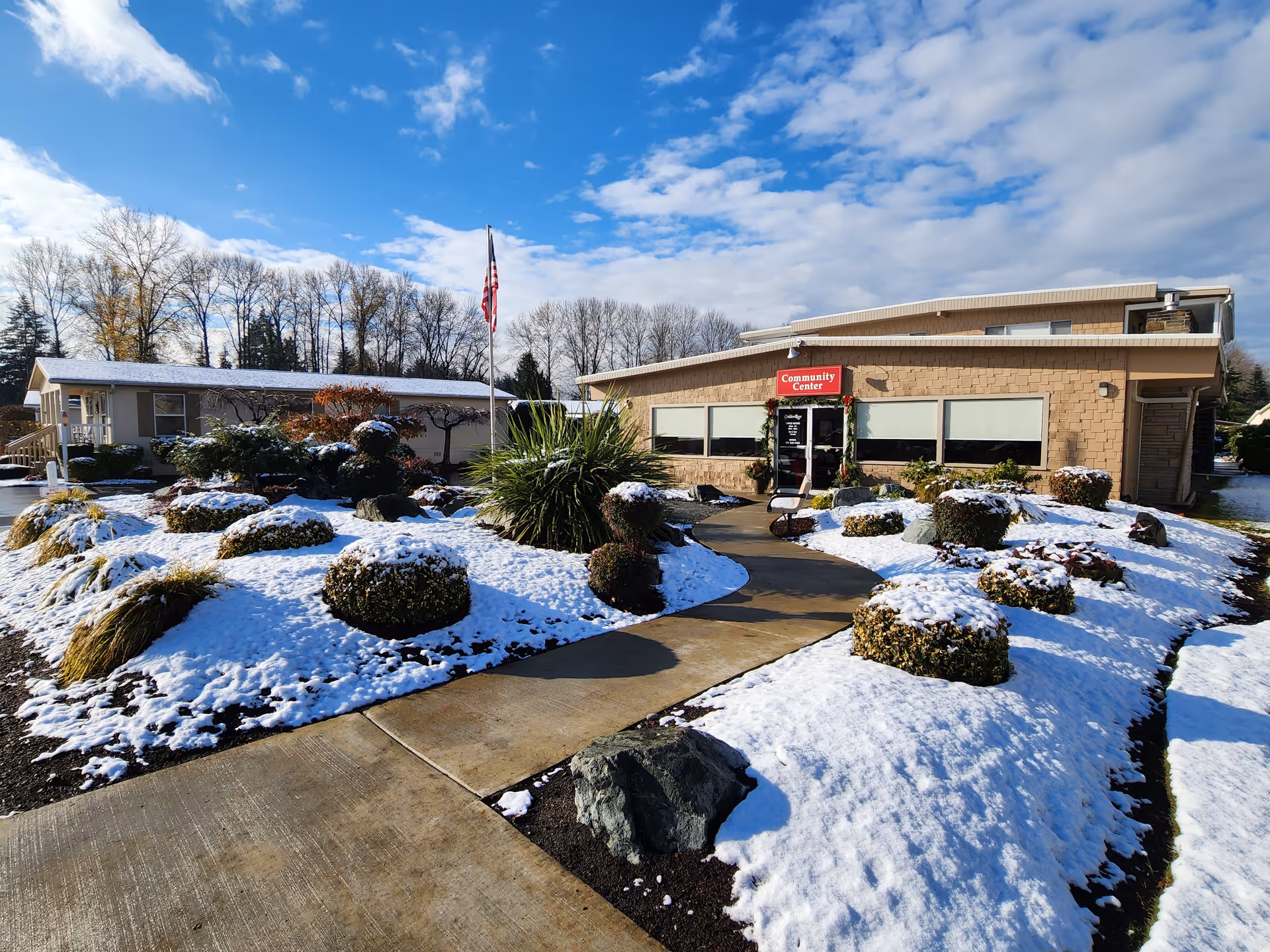 Exterior view of a community center building at Golden Rose 55+ Community near Tacoma, Washington, with a snow-covered landscaped garden and a clear blue sky with some clouds.