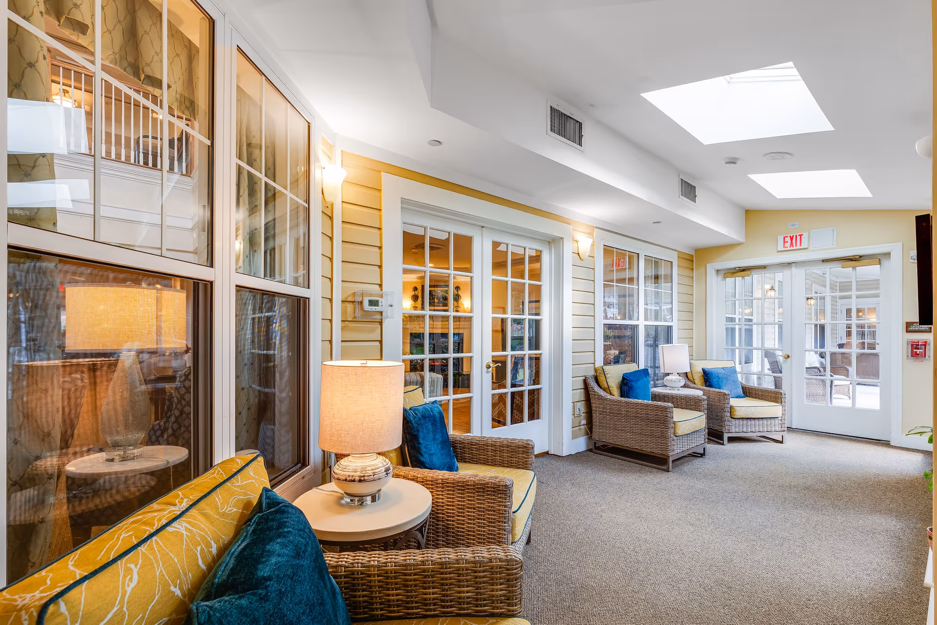 A bright and cozy indoor seating area with wicker armchairs featuring yellow cushions and blue pillows, a small round table with a lamp, large windows, glass doors, and skylights allowing natural light to fill the space.