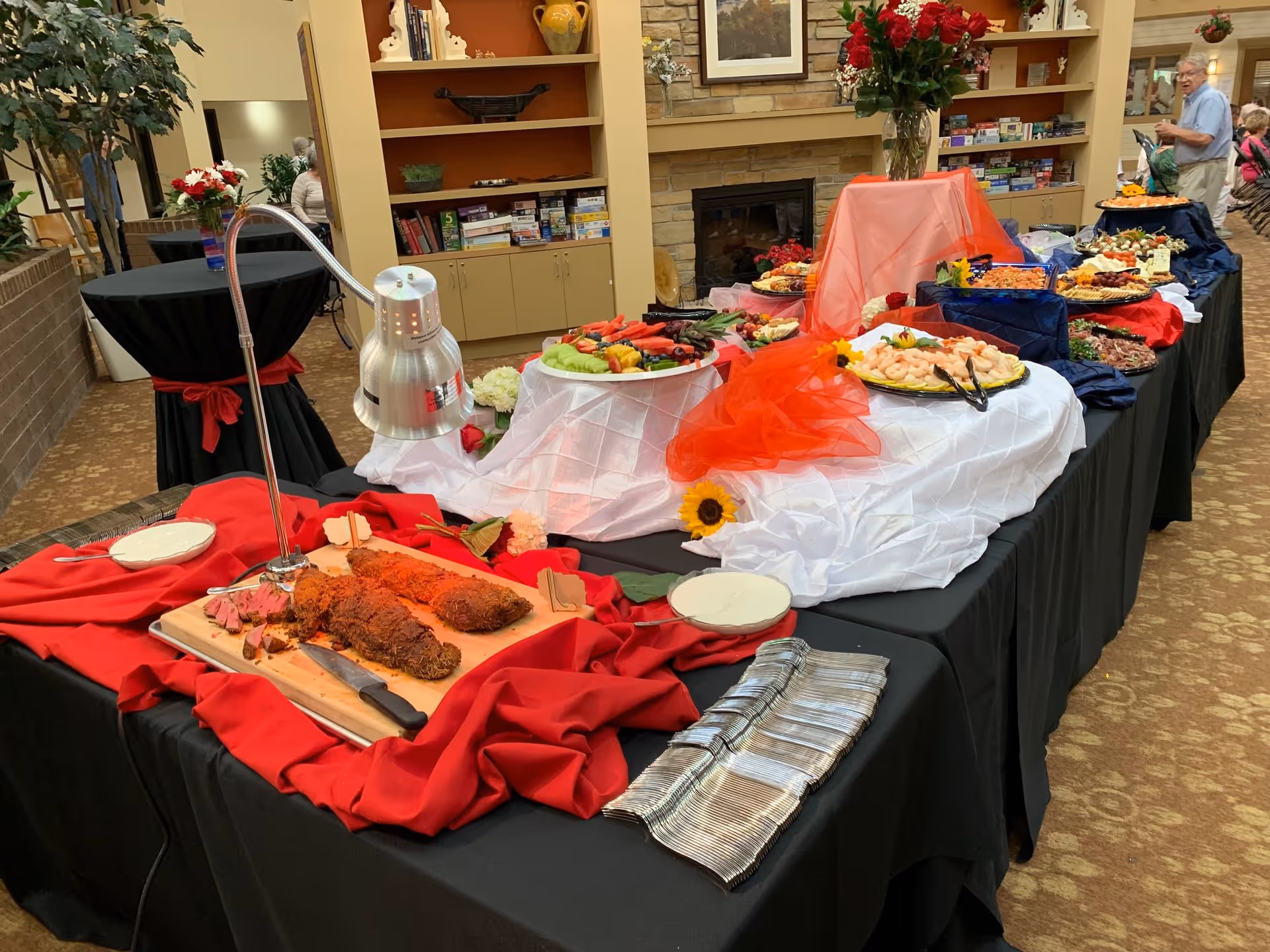 Buffet table set up with various foods including sliced meat, shrimp, fruit, and cheese platters, decorated with red and white cloths and flowers in a common area with bookshelves and a fireplace in the background. Several people are visible in the background.