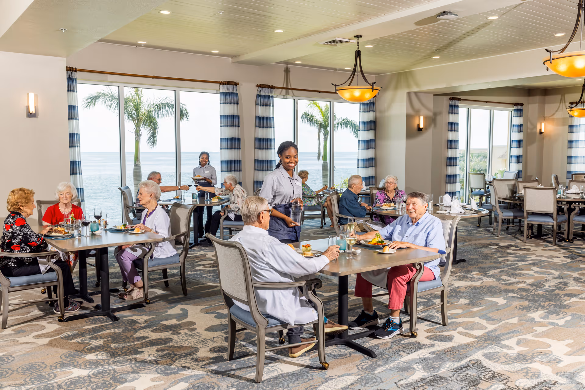 A bright dining room with large windows showing palm trees and a waterfront view. Several elderly people are seated at tables enjoying meals, while two staff members are serving food and drinks. The room has patterned carpet, light-colored walls, and blue-and-white striped curtains.