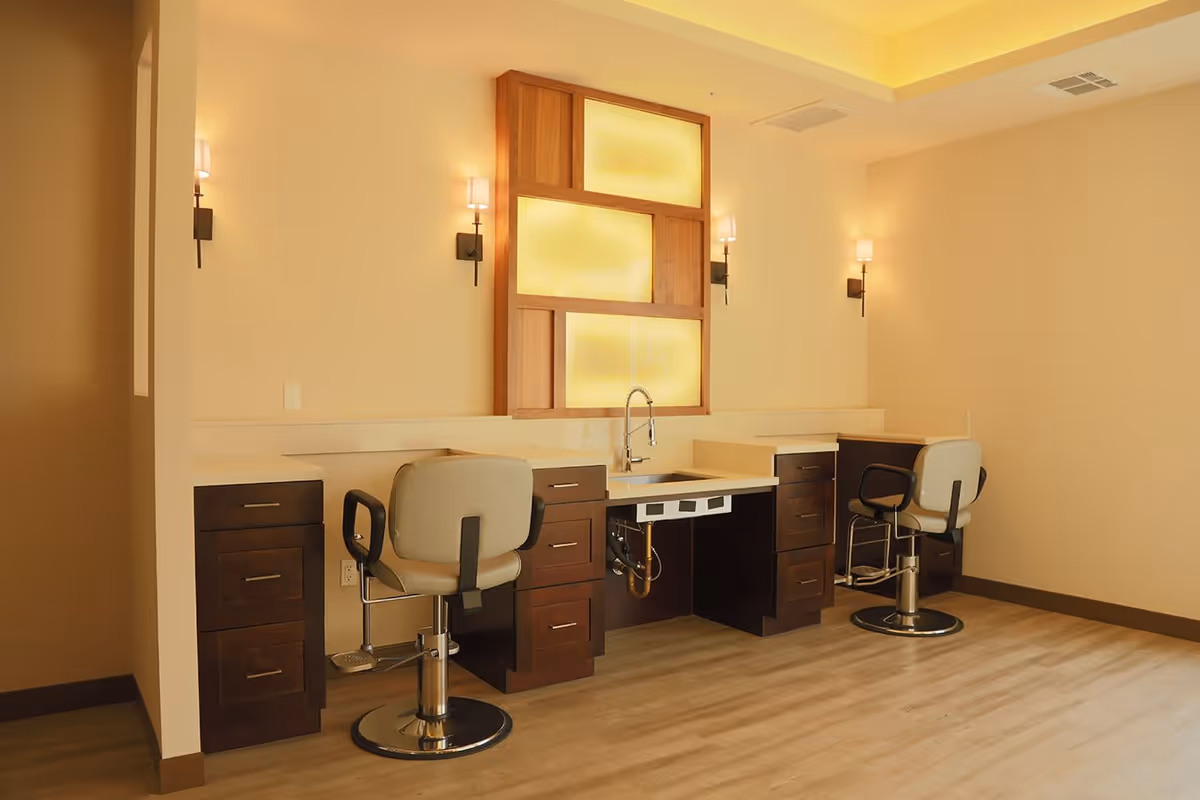 Interior view of a room with two salon-style chairs positioned in front of a counter with a sink in the middle. The counter has dark wood cabinets and drawers, and the wall behind features a decorative wooden panel with frosted glass. The room is softly lit with wall sconces and has light-colored walls and flooring.