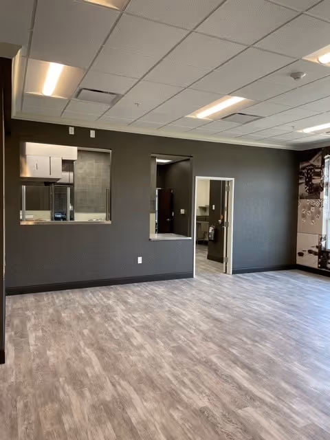 An empty interior room with gray walls and wood-patterned flooring. The room has a drop ceiling with fluorescent lights. There are two rectangular openings in the wall, one window-like and one door-like, revealing a kitchen area with stainless steel appliances and a hallway beyond. The right side of the image shows part of a wall with a black and white mural or photo collage.
