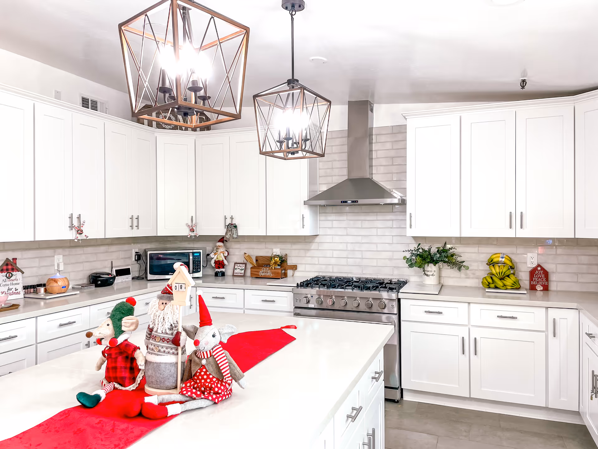 Bright modern kitchen with white cabinets, a large island countertop, stainless steel stove and range hood, and two geometric pendant lights. The island is decorated with festive holiday plush figures and a red runner. Various kitchen items and holiday decorations are placed on the countertops.