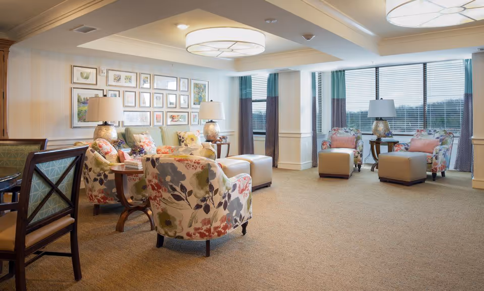 A bright and spacious living room area with floral upholstered armchairs, beige ottomans, side tables with lamps, and a wall decorated with framed artwork. Large windows with teal and purple curtains allow natural light to fill the room.