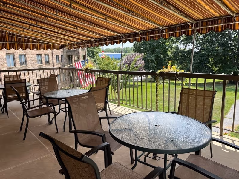 Covered outdoor balcony with round glass-top tables and chairs under a striped awning overlooking a grassy lawn.