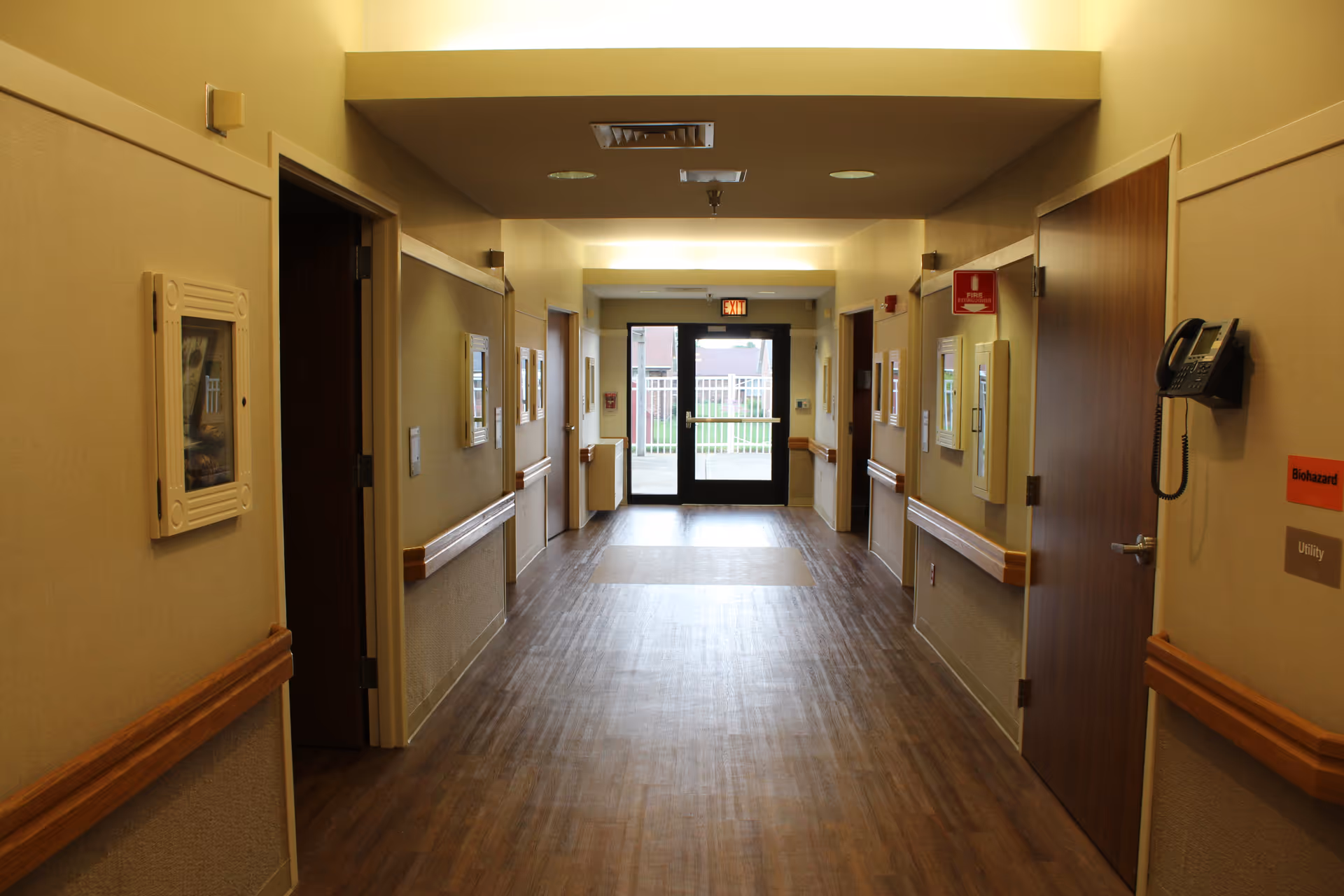 A well-lit hallway in a senior living facility with wooden floors, beige walls, handrails on both sides, several closed doors, a wall-mounted telephone, and an exit door with glass panels at the end.
