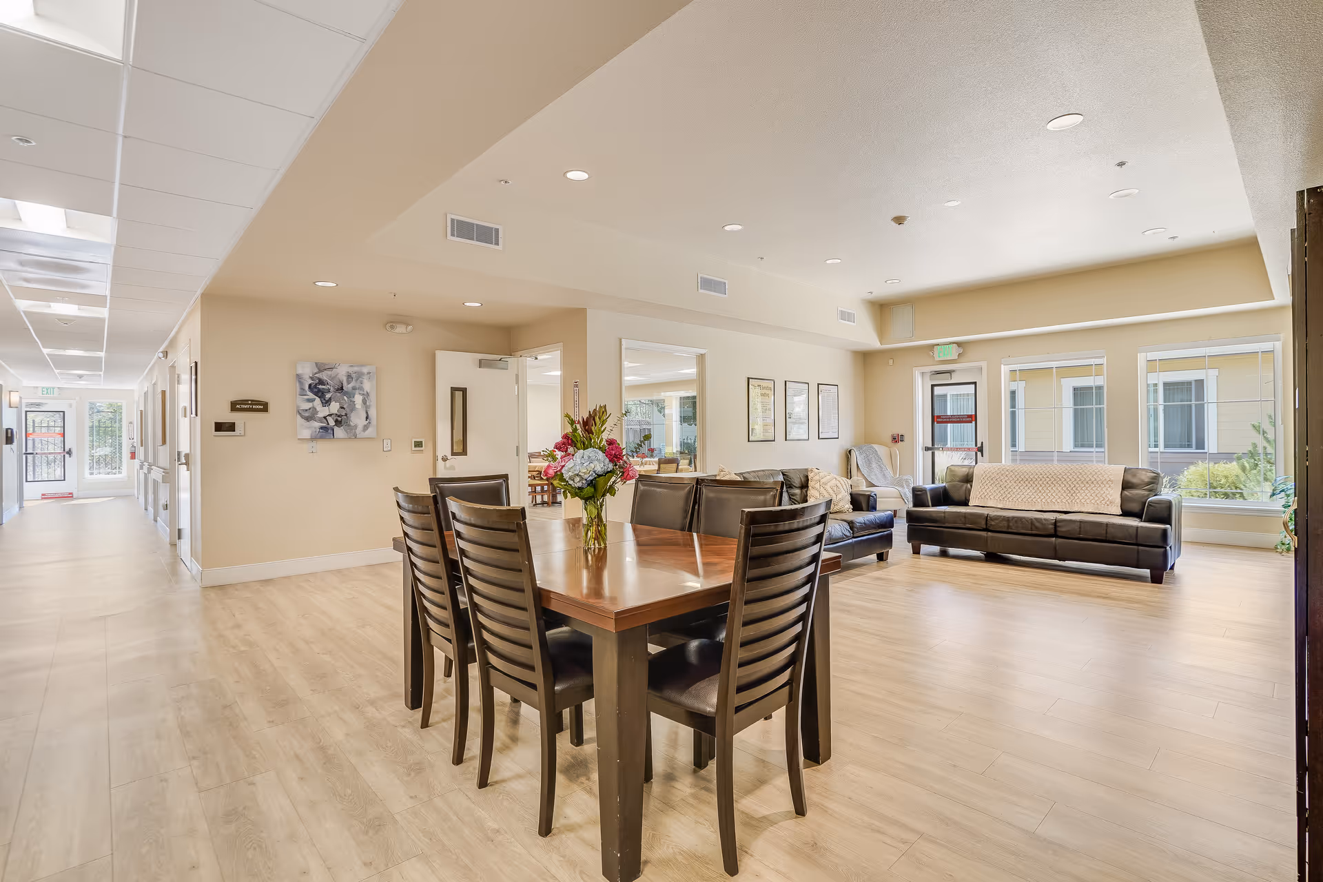 Bright senior living common area with a wooden dining table and chairs and sofas by large windows with a floral centerpiece on the table.