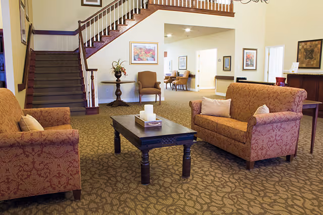 A cozy living room area in a senior living facility with two patterned armchairs and a matching loveseat arranged around a dark wooden coffee table with candles. In the background, there is a staircase with wooden banisters, a small round table with a plant, and framed artwork on the walls. The carpet has a leaf pattern and the walls are painted light yellow.