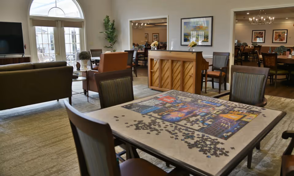 Communal activity room with a puzzle-covered table, chairs, sofa seating, a piano, and a dining area visible through doorways.